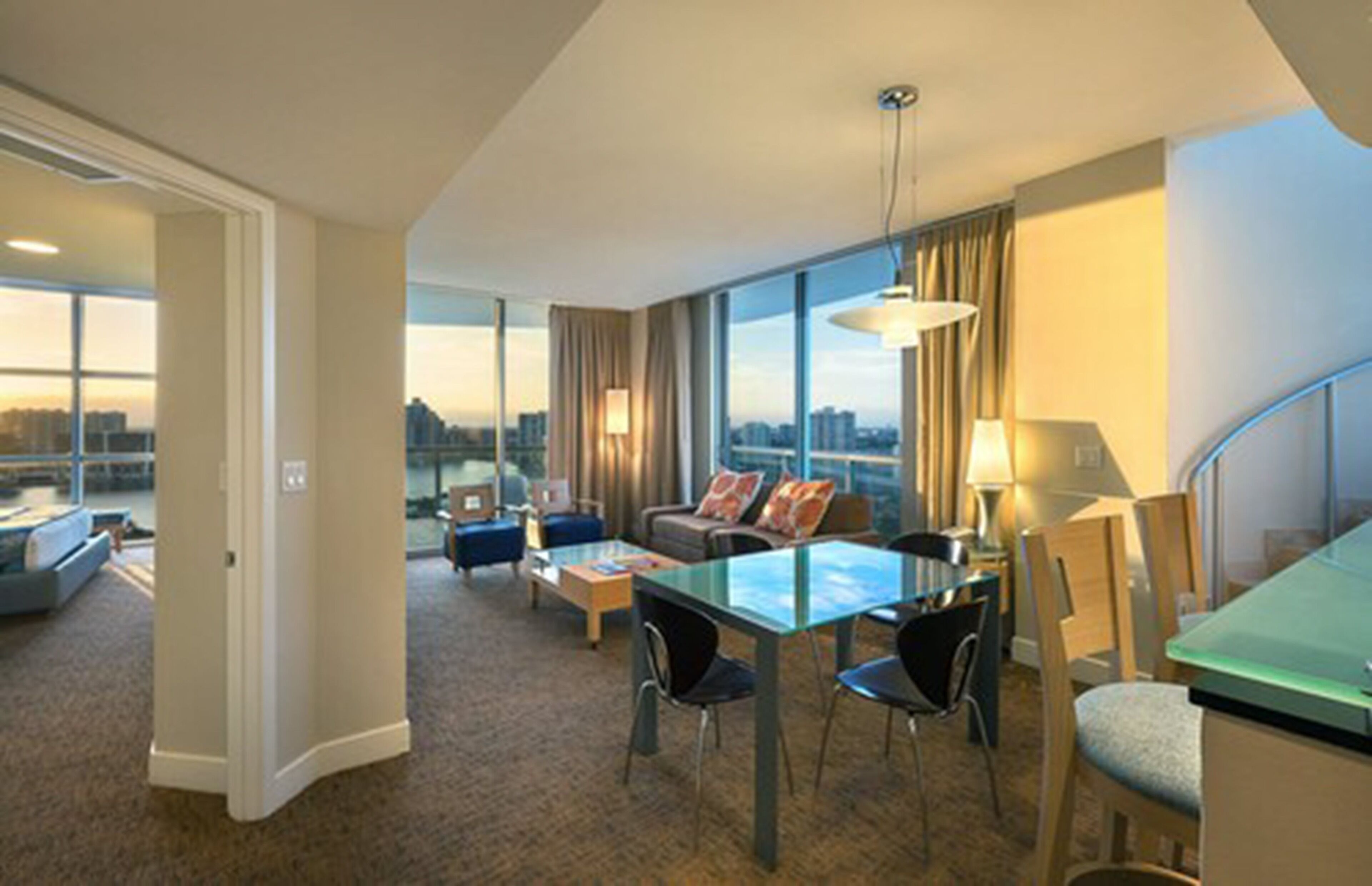 Una habitación de hotel moderna con zona de estar, mesa de comedor y grandes ventanales con vista a la ciudad.