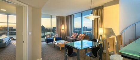Una habitación de hotel moderna con zona de estar, mesa de comedor y grandes ventanales con vista a la ciudad.
