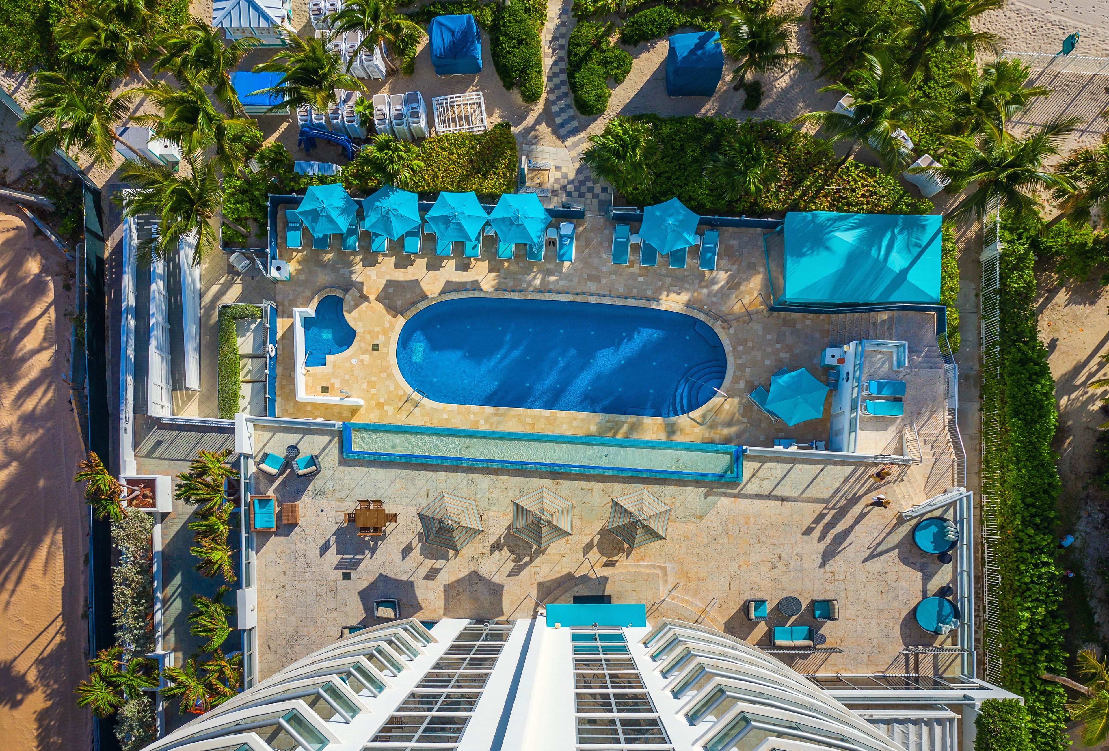 An aerial view of a resort with a central swimming pool, sun loungers, and palm trees.