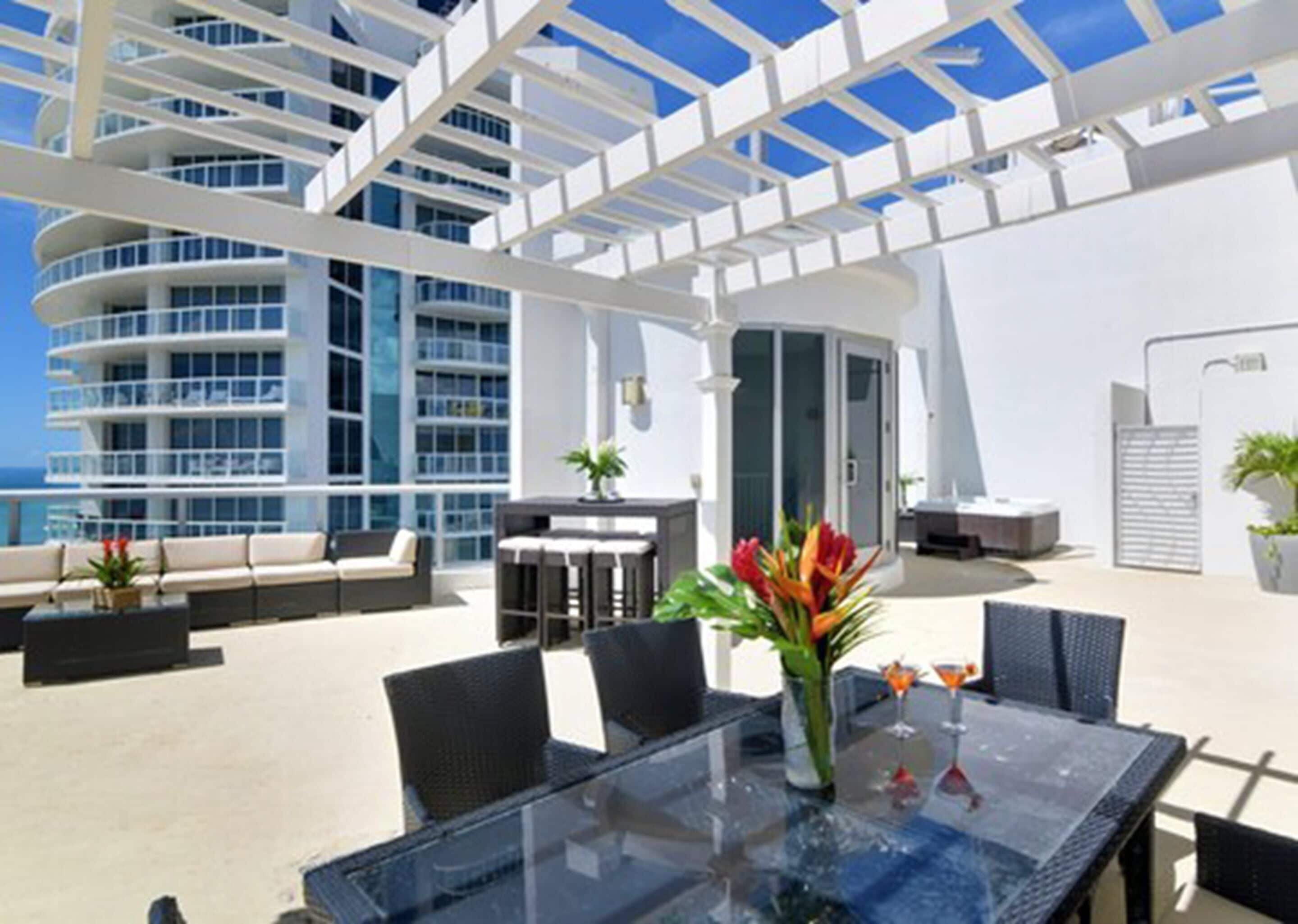 A sunlit outdoor area with a glass table, chairs, and a sofa. There is a view of a modern building with balconies and a clear sky.