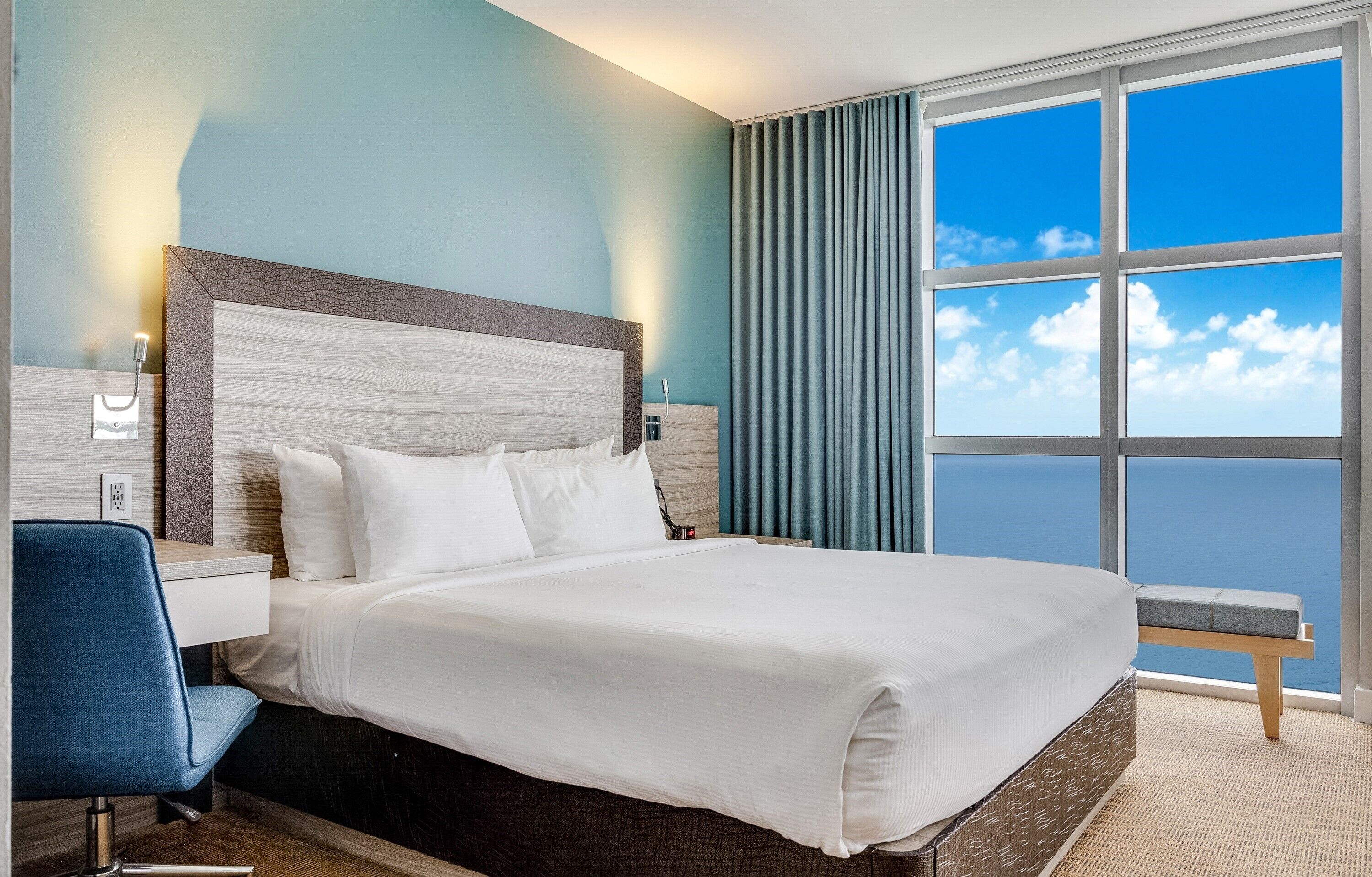 A hotel room with a large bed, a desk, a chair, and a view of the ocean through a window.