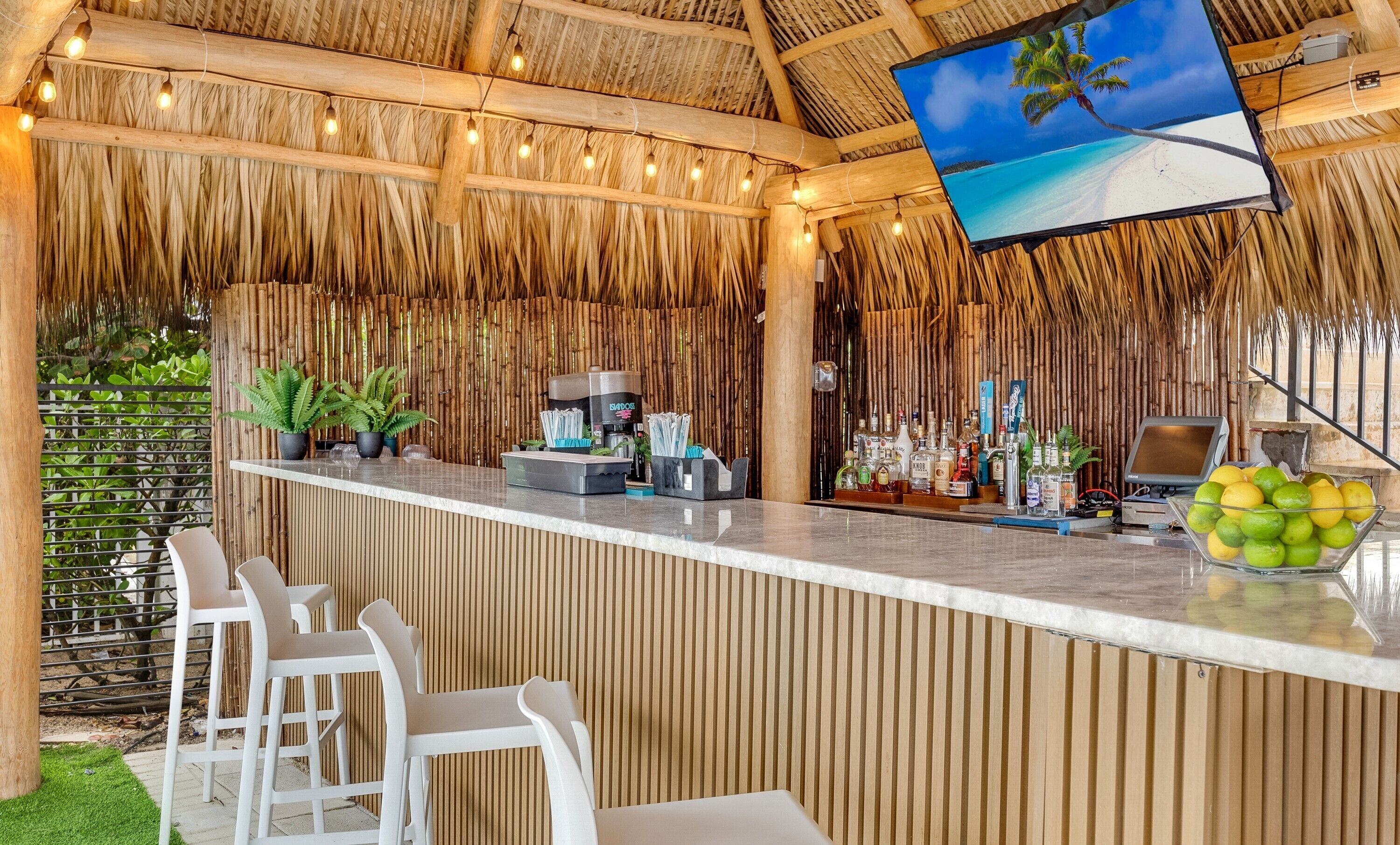 Una zona de bar con techo de paja, un televisor de pantalla plana mostrando una escena de playa y una variedad de botellas y vasos sobre la barra.