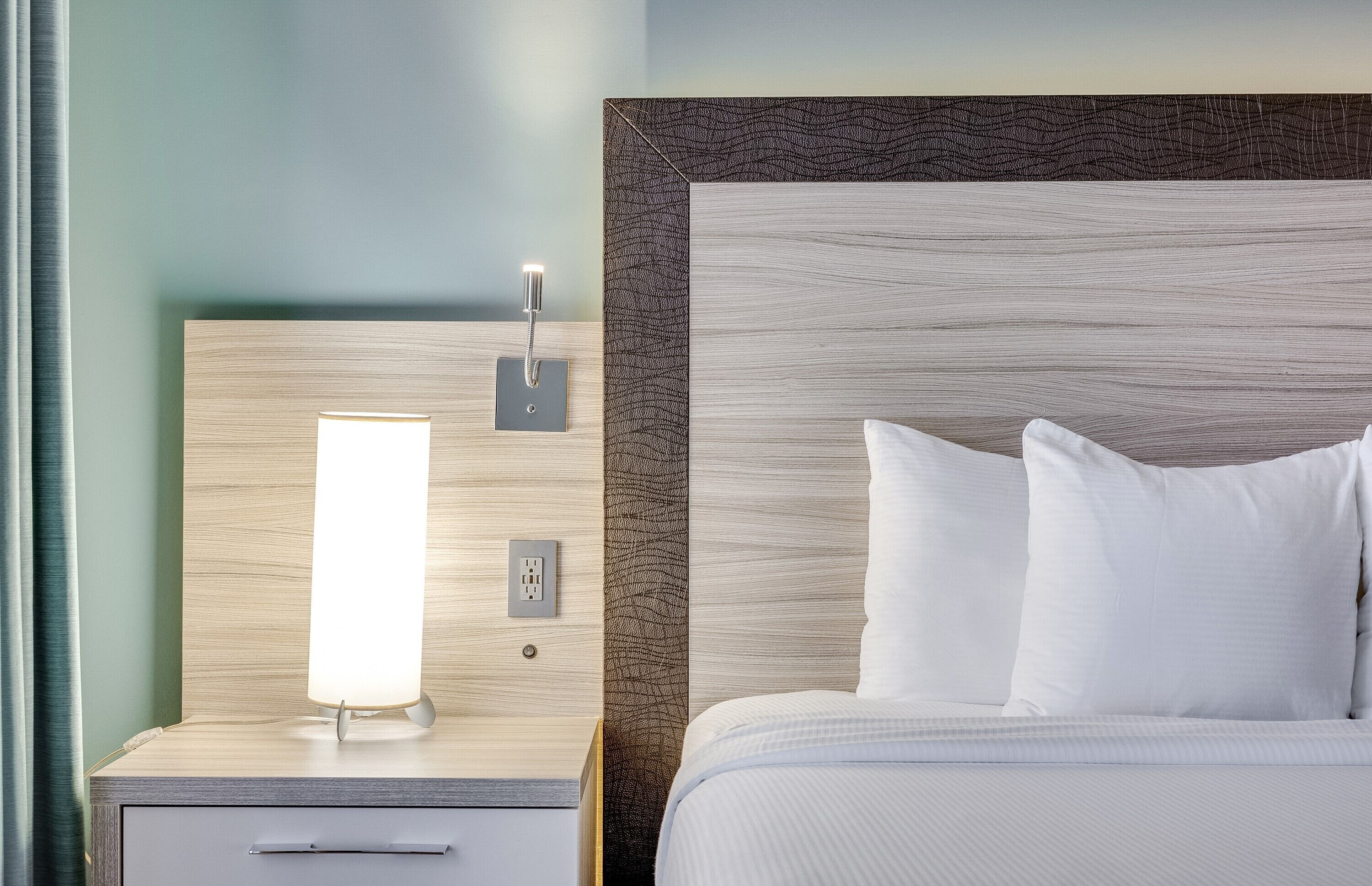 A hotel room with a wooden headboard, a bedside table with a lamp, and a bed with white bedding.