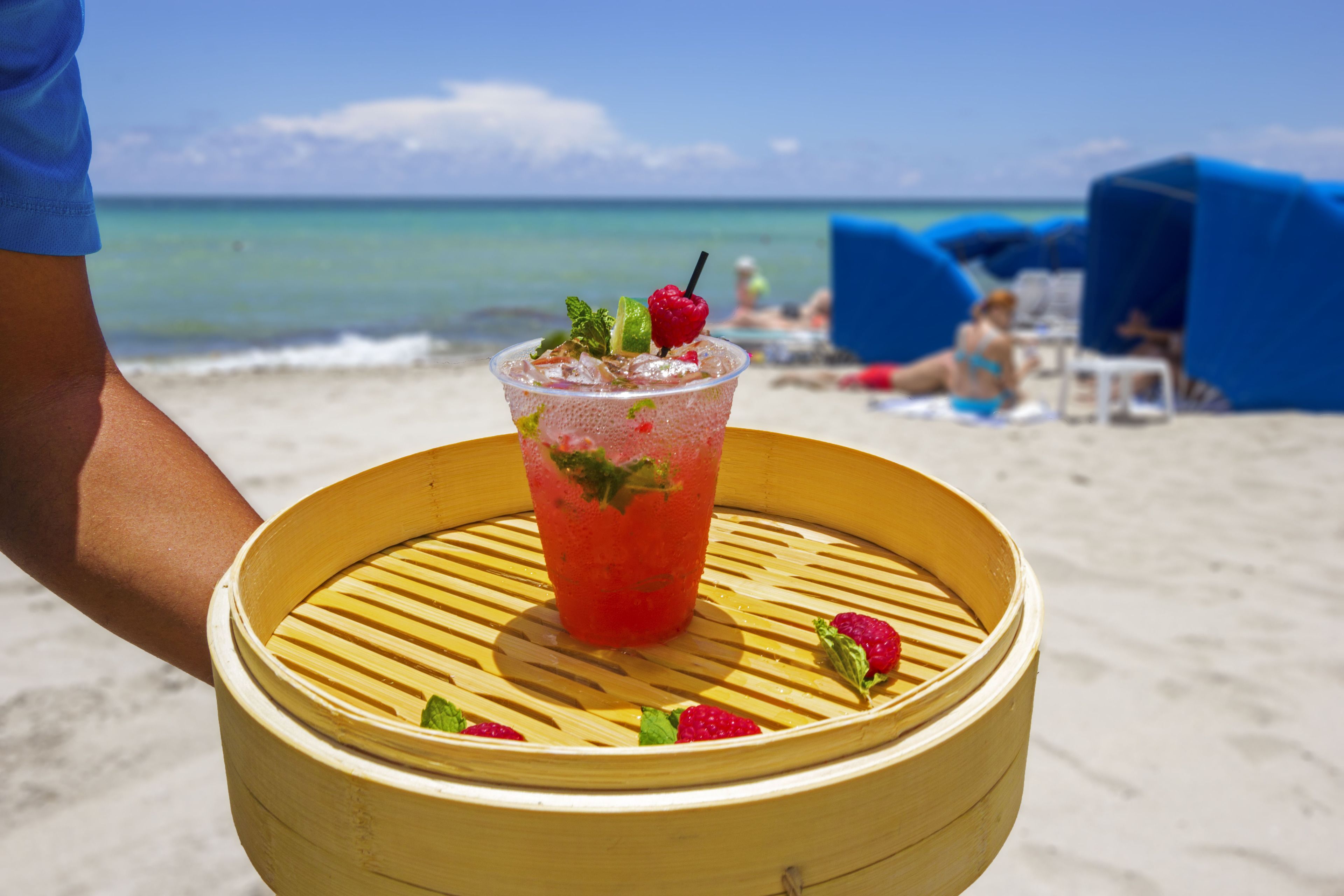A drink in a bamboo steamer on a beach with umbrellas and people in the background.