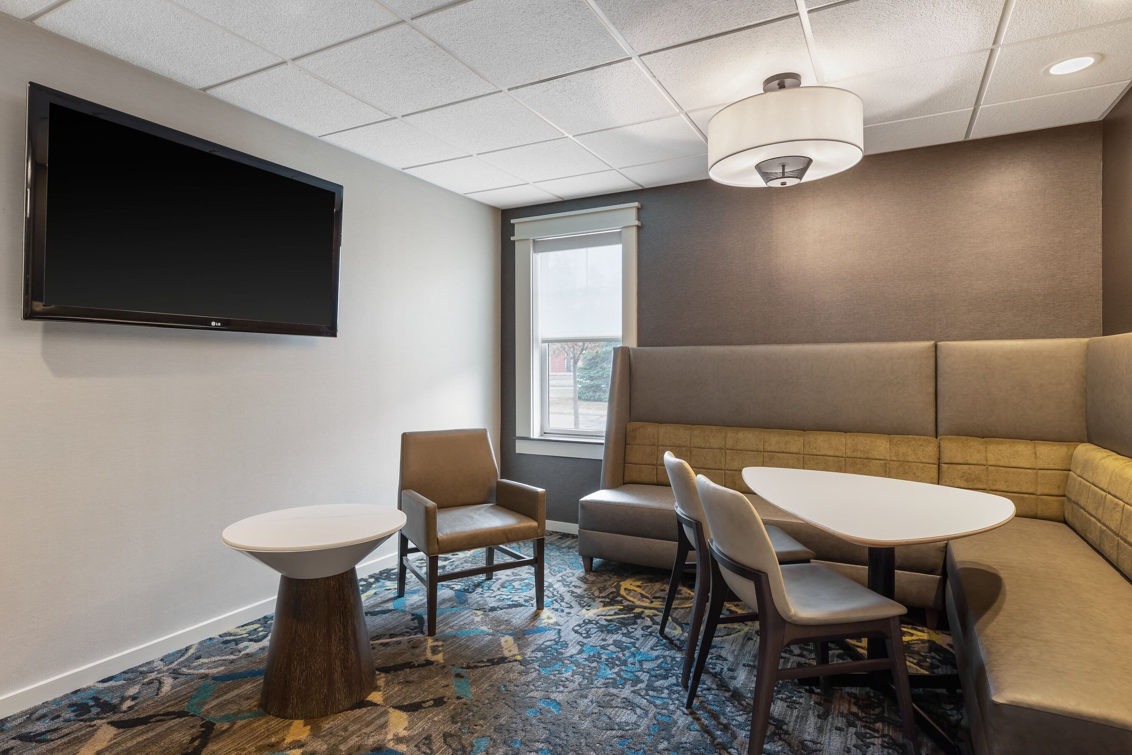 A conference room with a large flat-screen TV, a round table, two chairs, a sofa, and a patterned carpet.