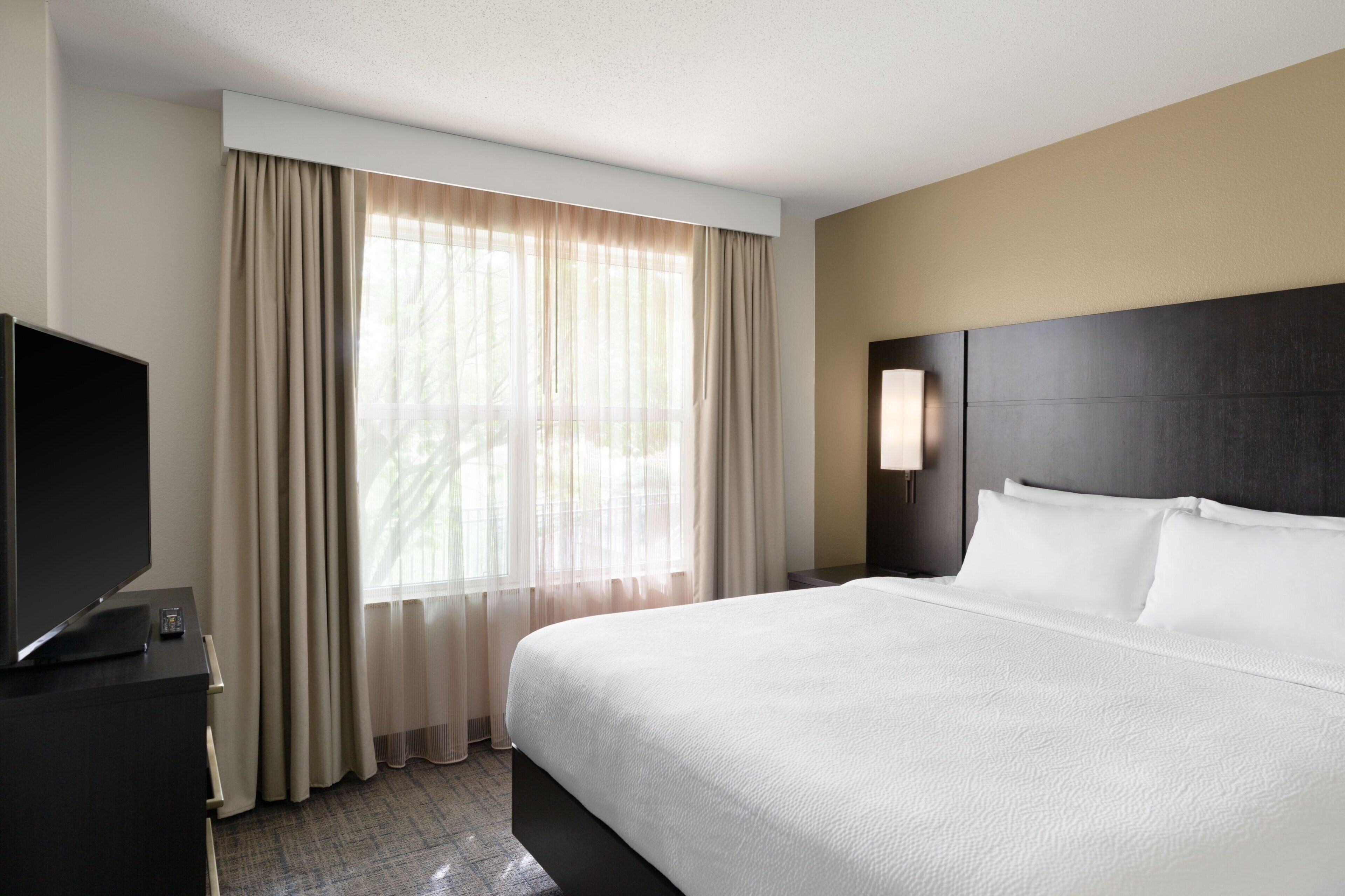 Una habitación de hotel con una cama grande, un televisor de pantalla plana sobre una cómoda oscura, y una ventana con cortinas translúcidas.