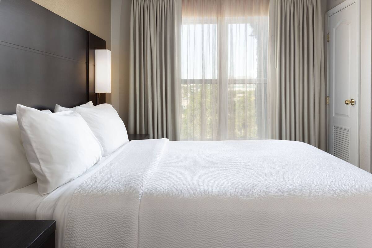 A hotel room with a large bed, a nightstand, a lamp, and a view of the outdoors through sheer curtains.