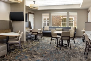 A modern hotel room with a large window, a sofa, a dining table, and chairs.