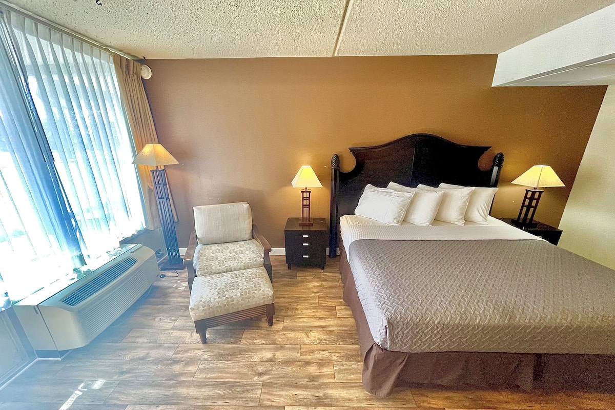Habitación de hotel con cama, una silla, dos mesitas de noche con lámparas, un calentador y una ventana con cortinas.