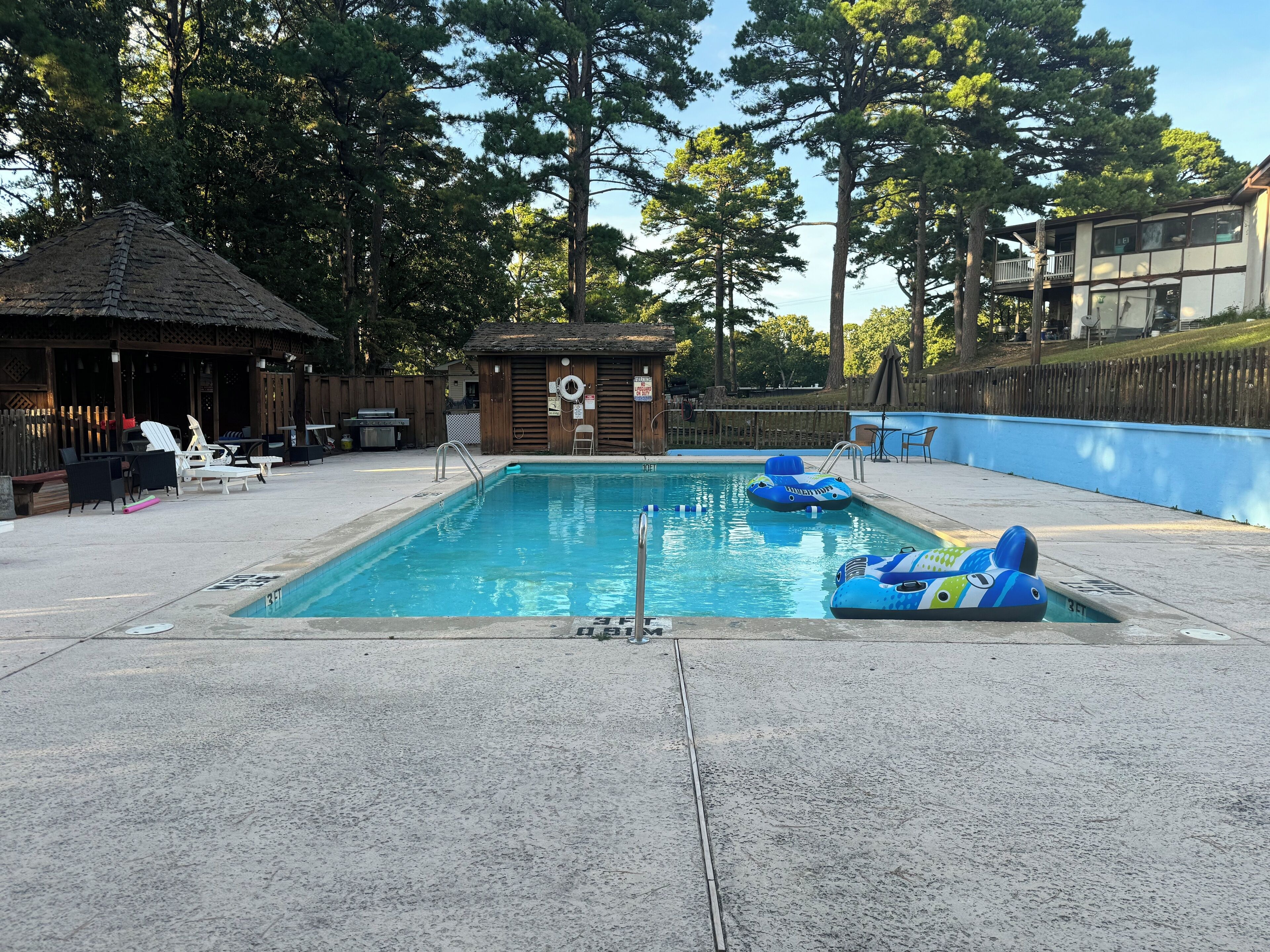 A swimming pool with lounge chairs and inflatable toys, surrounded by trees and a wooden fence.