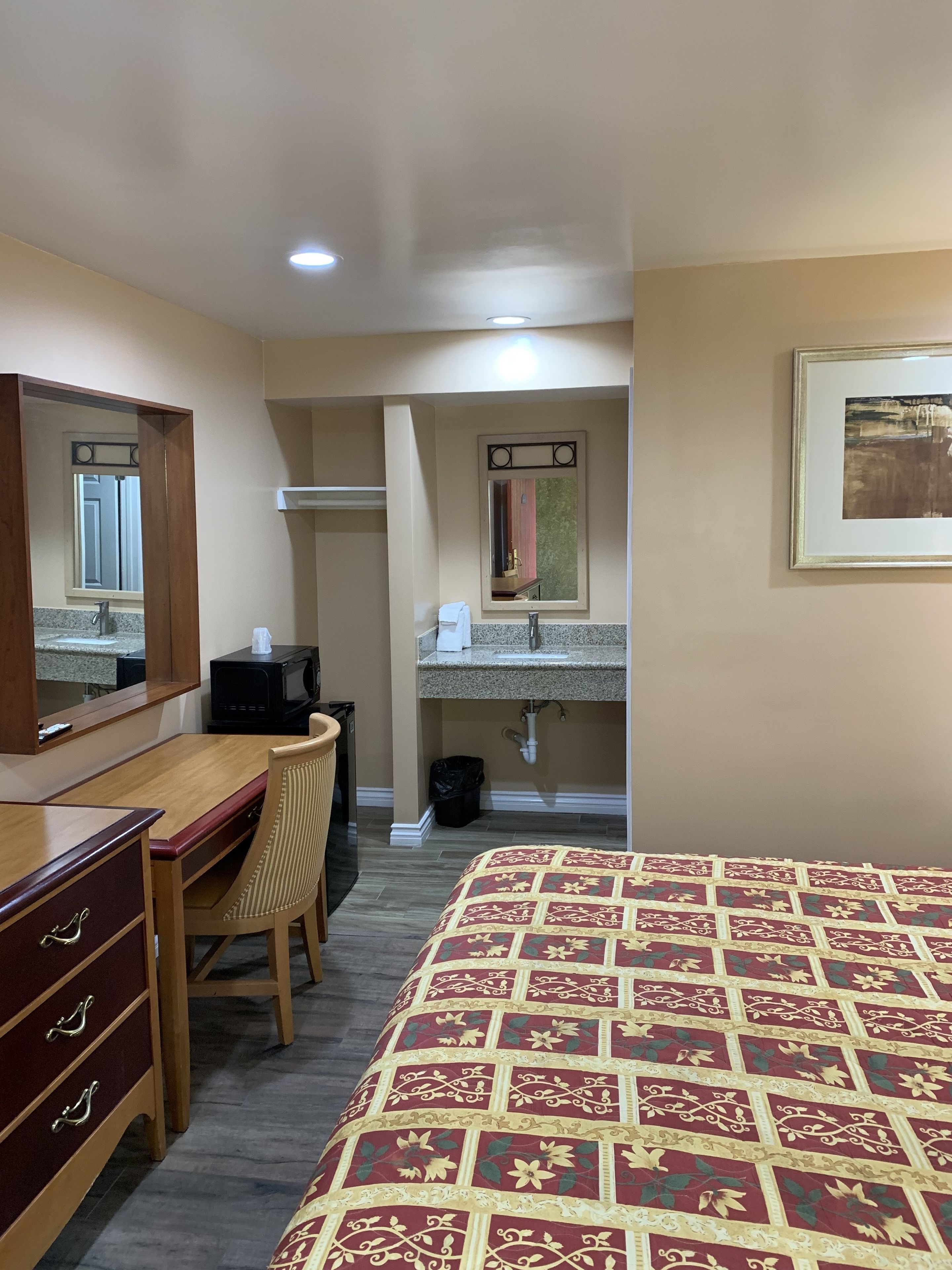 A hotel room with a wooden desk, a chair, a bed with a patterned bedspread, a bathroom with a sink and mirror, and a framed picture on the wall.