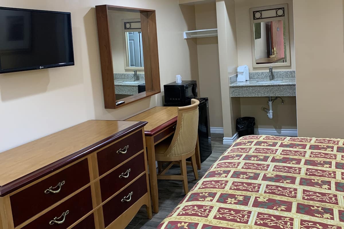 A hotel room with a bed, a desk with a chair, a television, a mirror, a microwave, and a sink with a towel.