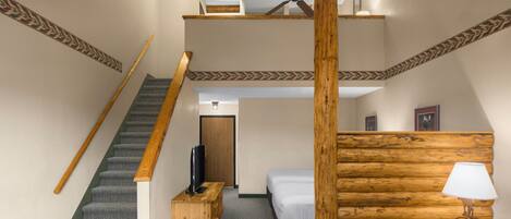 Una habitación de hotel con una escalera, una cama, cabecera de madera, una mesita de noche, una lámpara y un televisor.