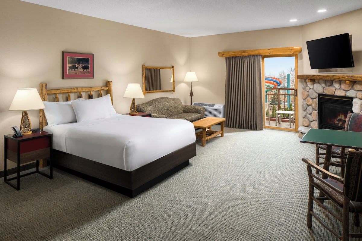 A hotel room with a large bed, two bedside tables with lamps, a television mounted above a fireplace, and a view of an outdoor area with a slide.