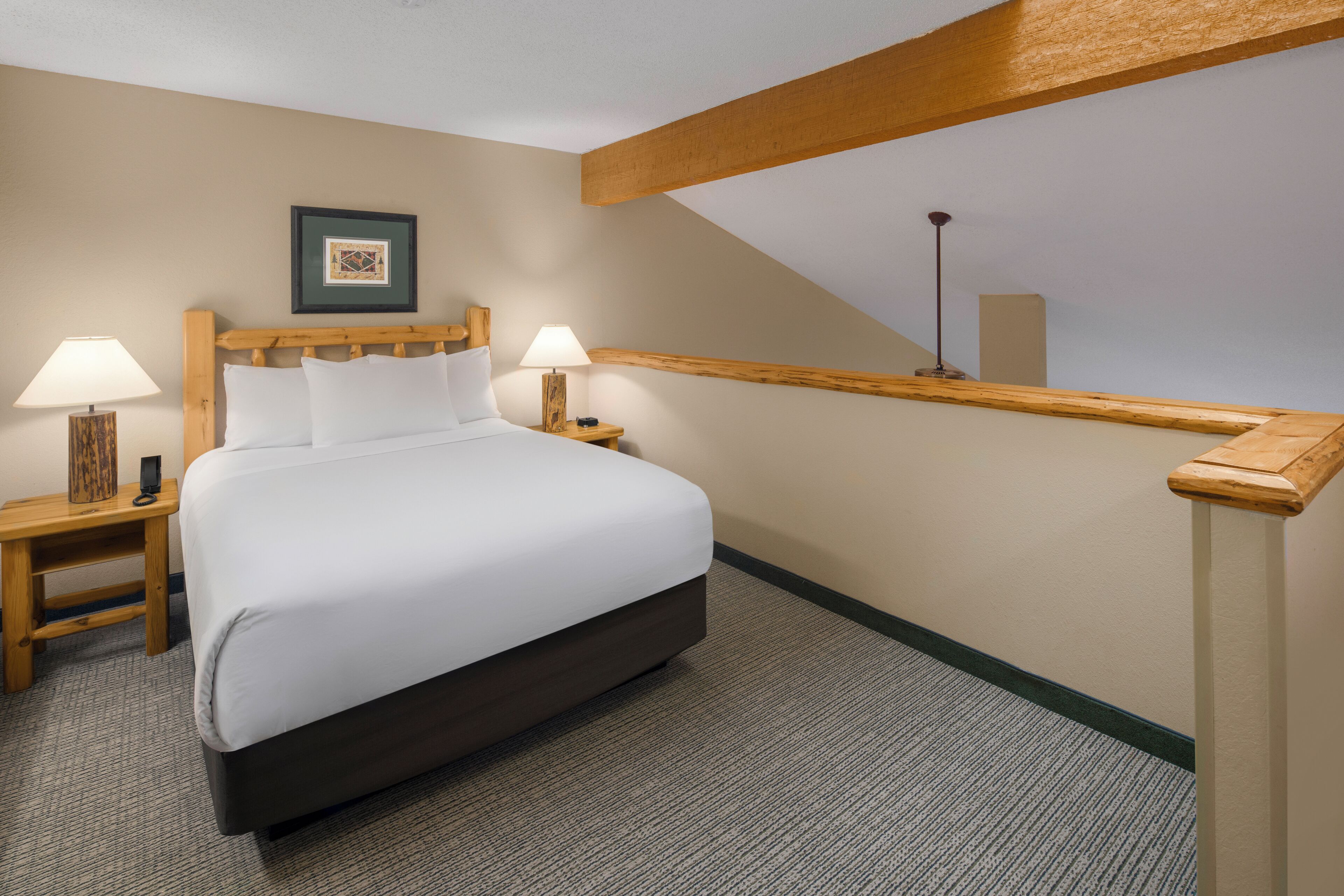 A hotel room with a large bed, two bedside tables with lamps, a framed picture on the wall, and a sloped ceiling with wooden beams.
