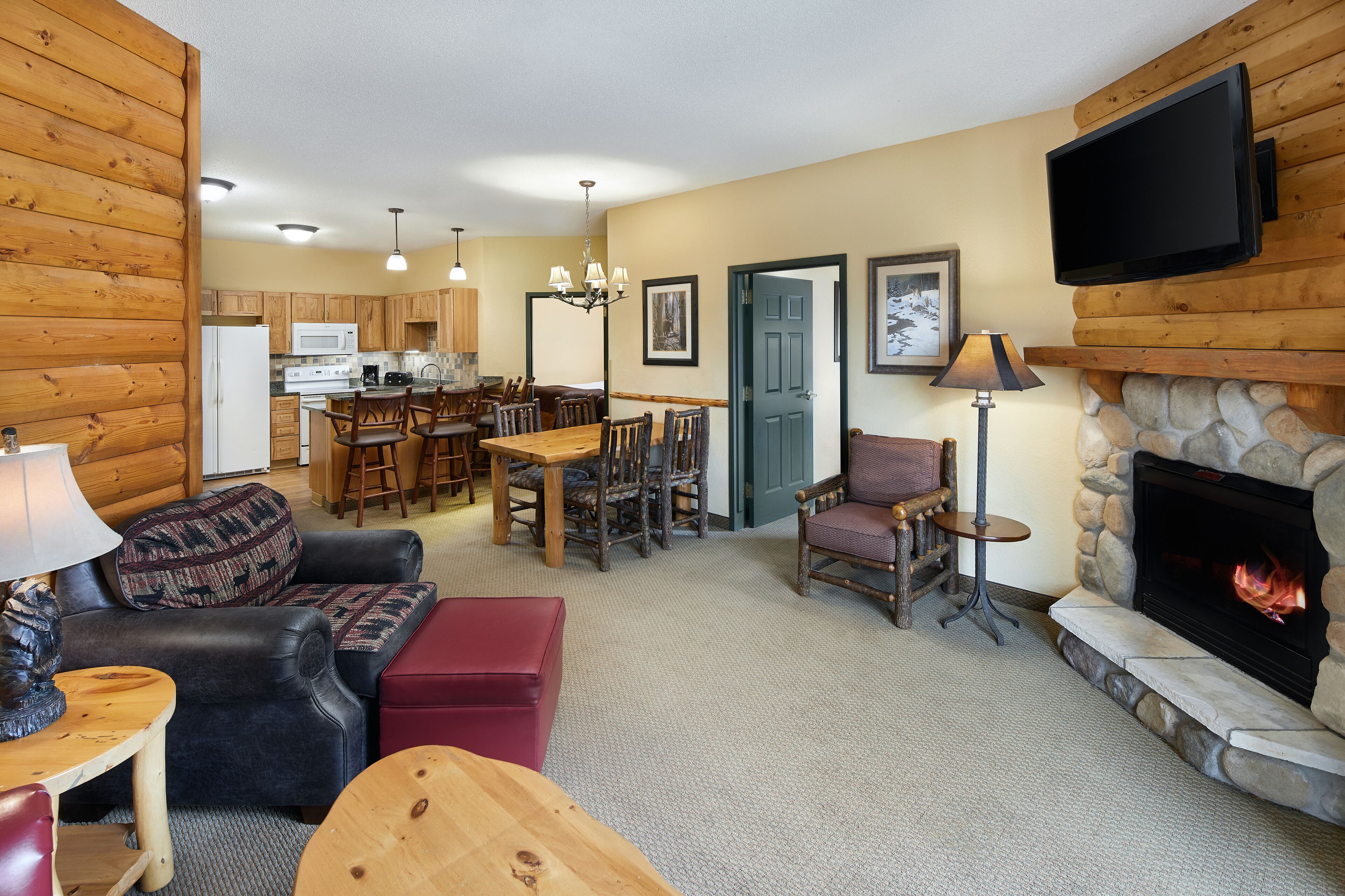 A cozy living room with a fireplace, wooden walls, a dining area, and a kitchen in the background.