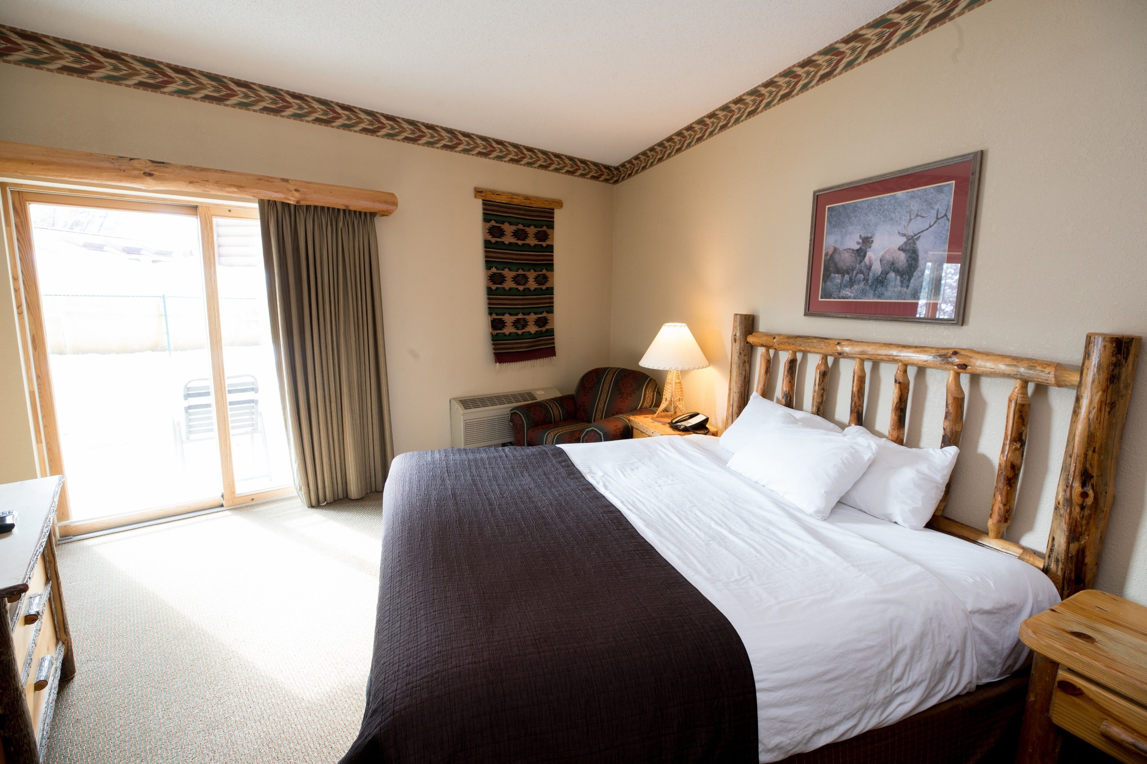 A hotel room with a large bed, a wooden headboard, a desk, a chair, a lamp, and a framed picture on the wall.
