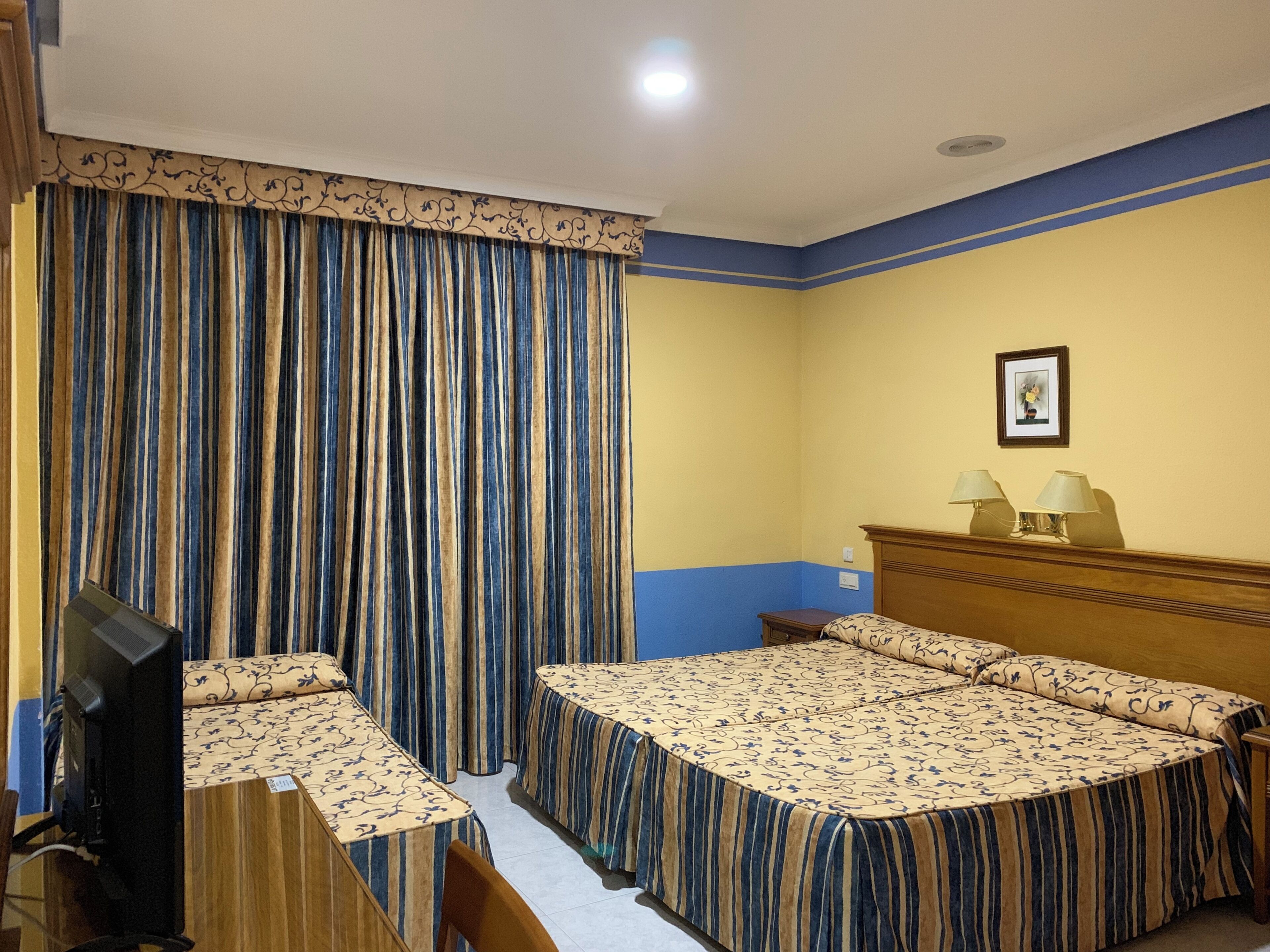 A hotel room with a bed, a desk, and a chair. There is a television on the desk. The room has a curtain and a picture on the wall.