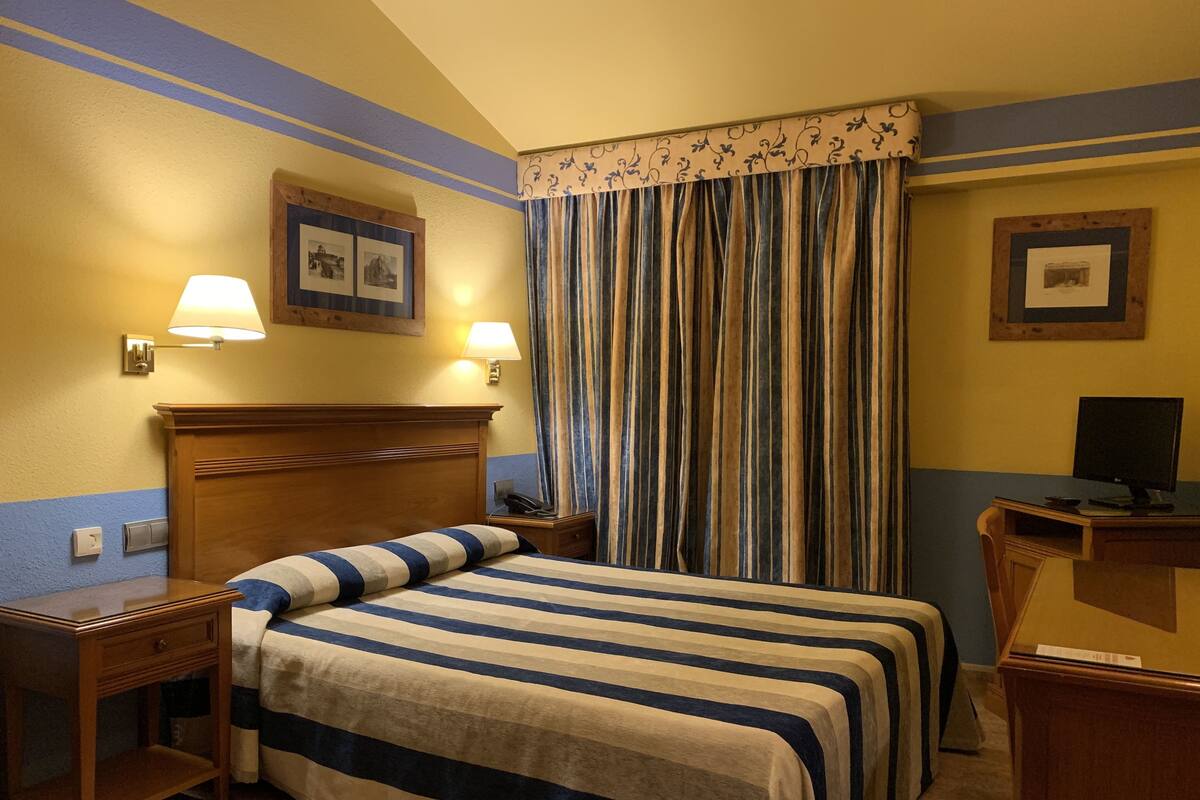 A hotel room with a double bed, two bedside tables, a desk with a computer, a lamp, and a curtain.