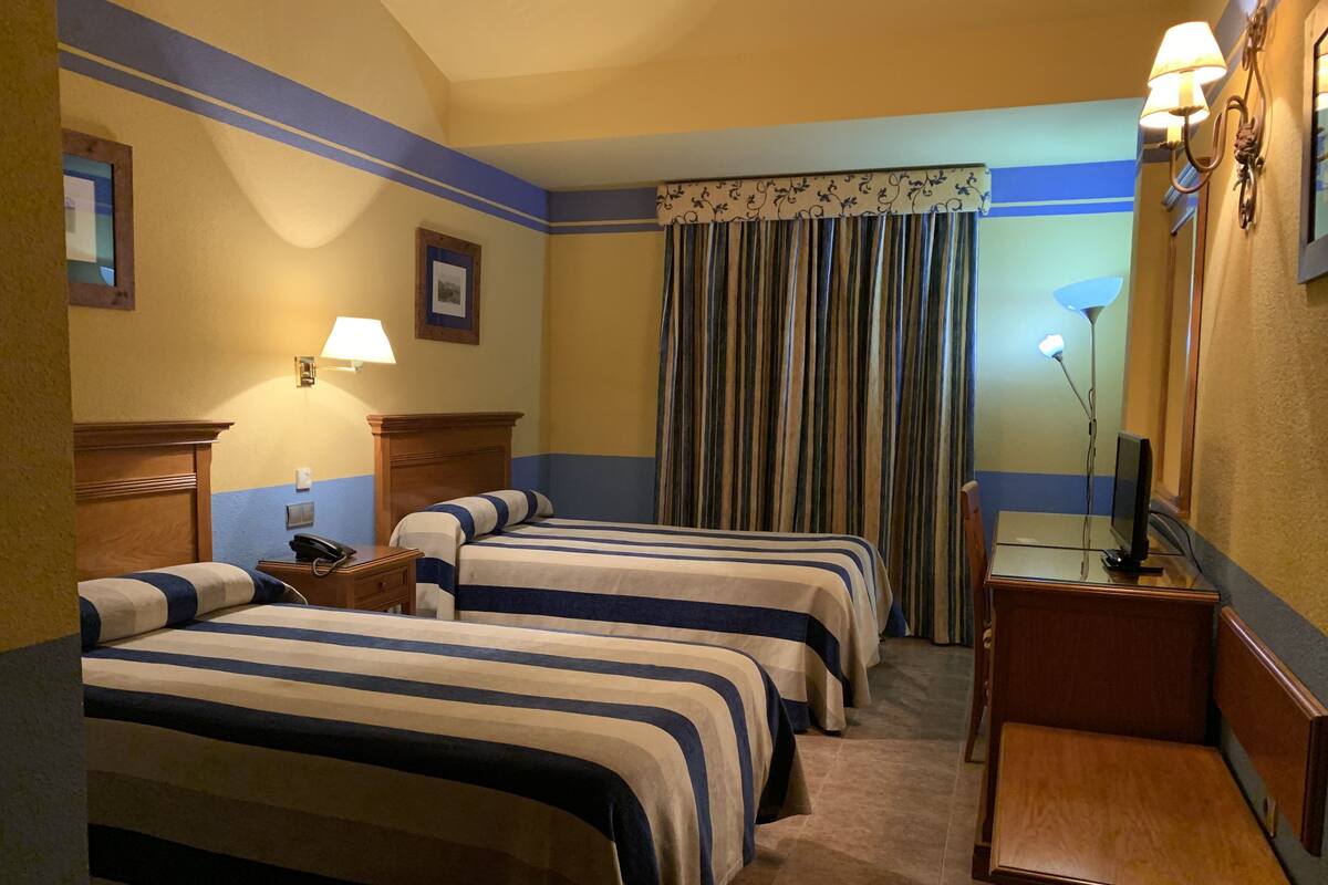 A hotel room with three beds, a desk, a chair, a lamp, a mirror, and a window with curtains.