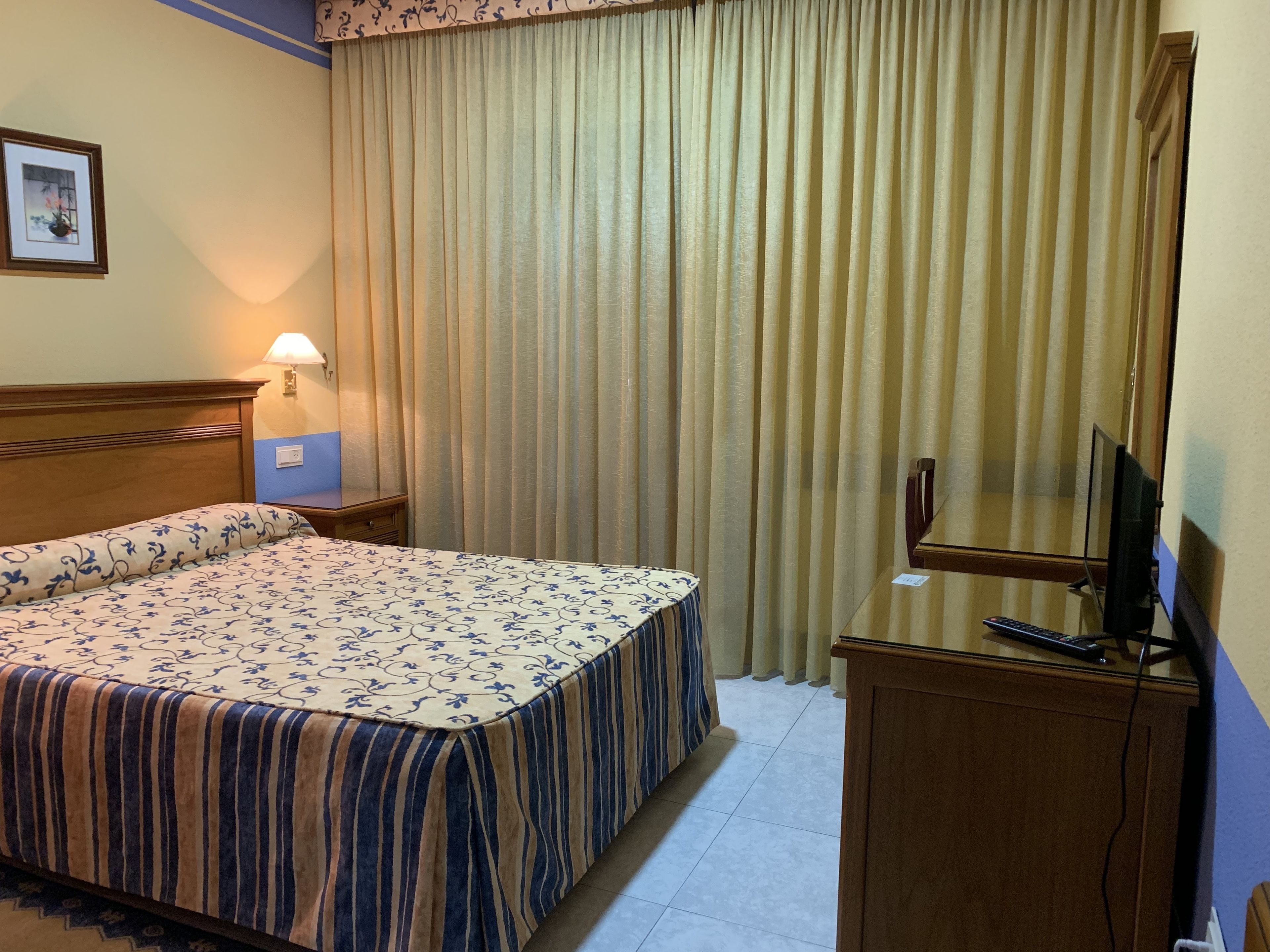 A hotel room with a bed, a desk with a television, a lamp, and a picture on the wall.