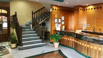 A hotel lobby with a marble floor, a reception desk, a staircase with ornate railings, and framed certificates on the wall.