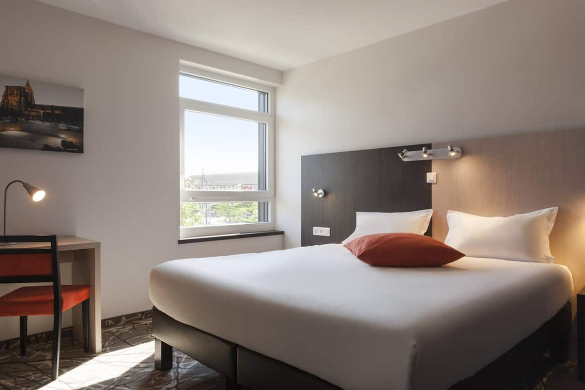 Une chambre d’hôtel avec un grand lit, un bureau, une chaise, une fenêtre et un tableau au mur.