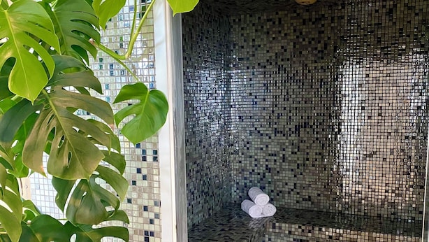 A room with mosaic tile walls and floor, a seating area with a small table and rolled towels, and a potted plant with large leaves. - The Originals Boutique, Hôtel d'Alsace, Strasbourg Sud (Illkirch-Graffenstaden)