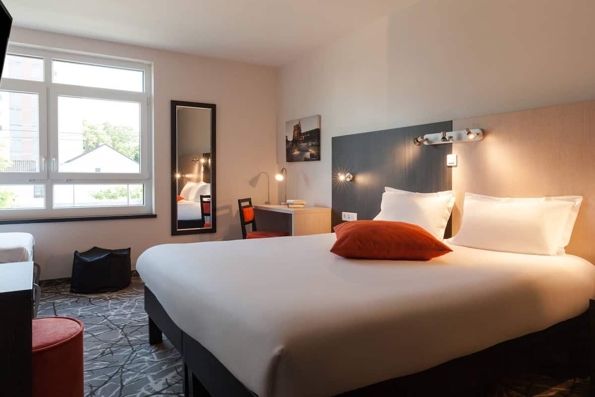 Une chambre d’hôtel avec un grand lit, un bureau, une chaise, un miroir et une fenêtre donnant sur les bâtiments environnants.