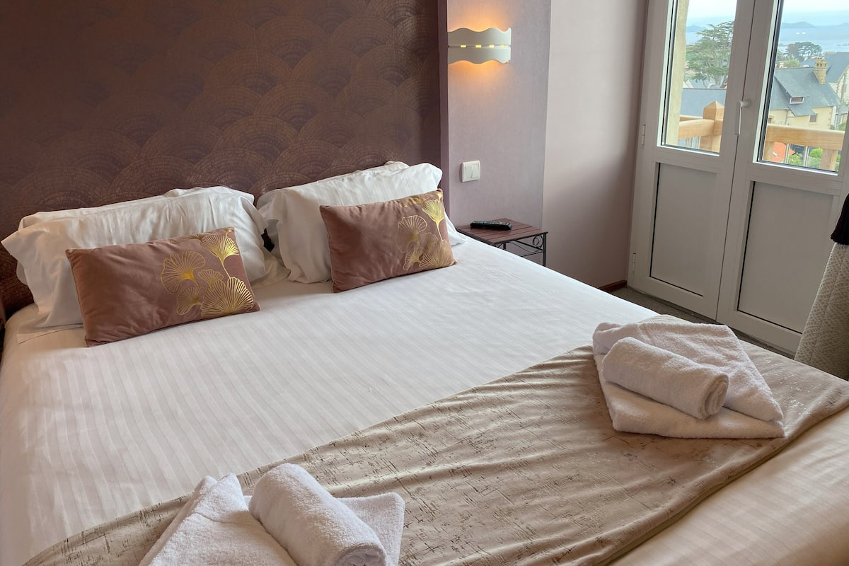 A hotel room with a large bed, two pillows, a nightstand, a remote control, and a window with a view of buildings and trees.