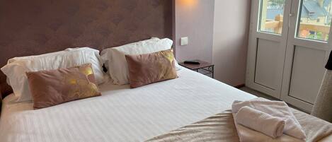 Habitación de hotel con una cama grande, dos almohadas, una mesita de noche, un control remoto y una ventana con vistas a edificios y árboles.