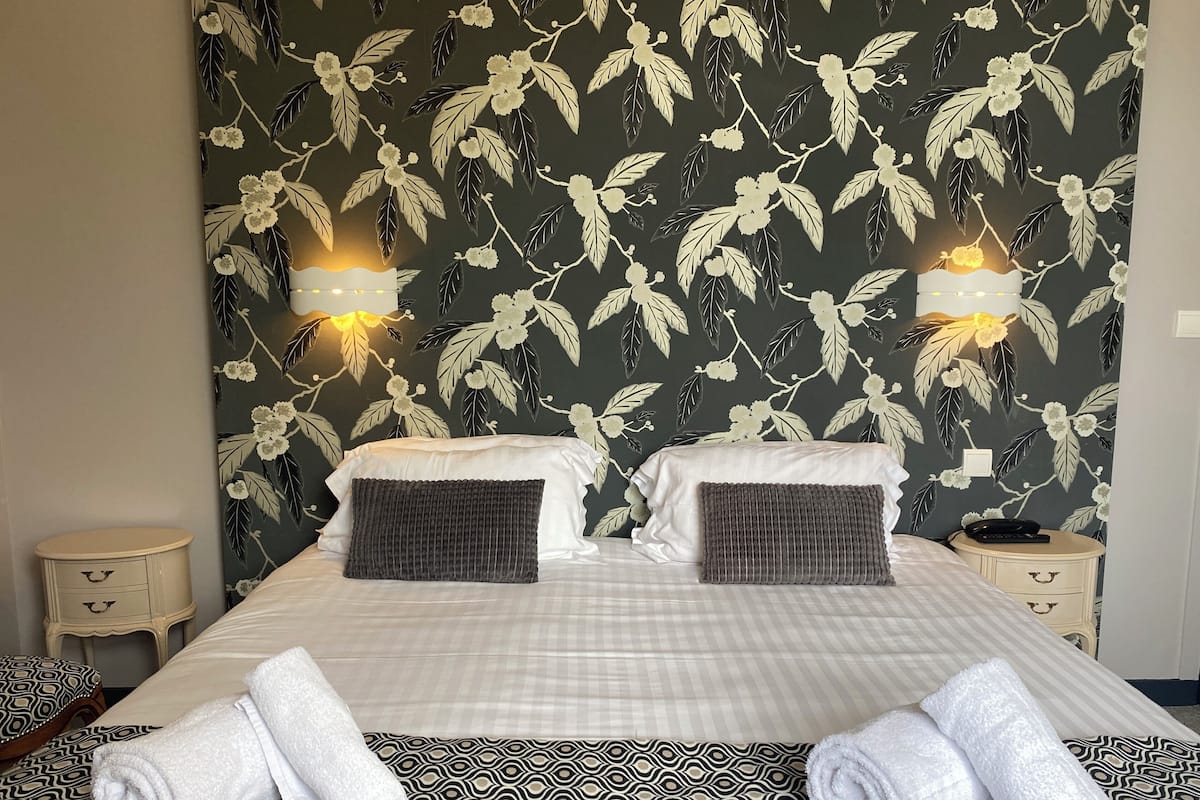 A hotel room with a large bed, two bedside tables, a wall with floral wallpaper, and two wall-mounted lamps.