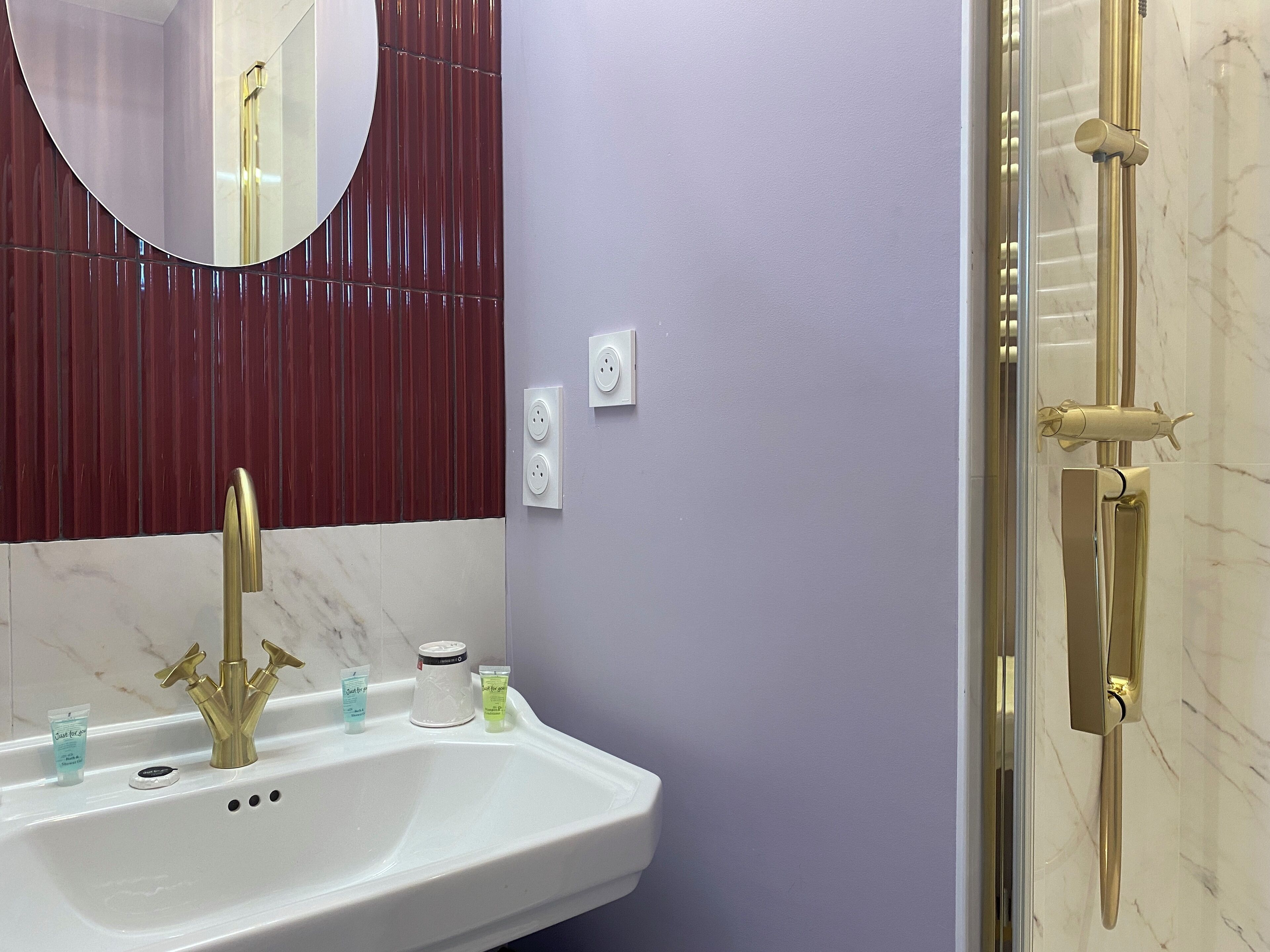 Une salle de bain avec un lavabo blanc, des robinets dorés, un miroir rond et une douche avec des accessoires dorés.