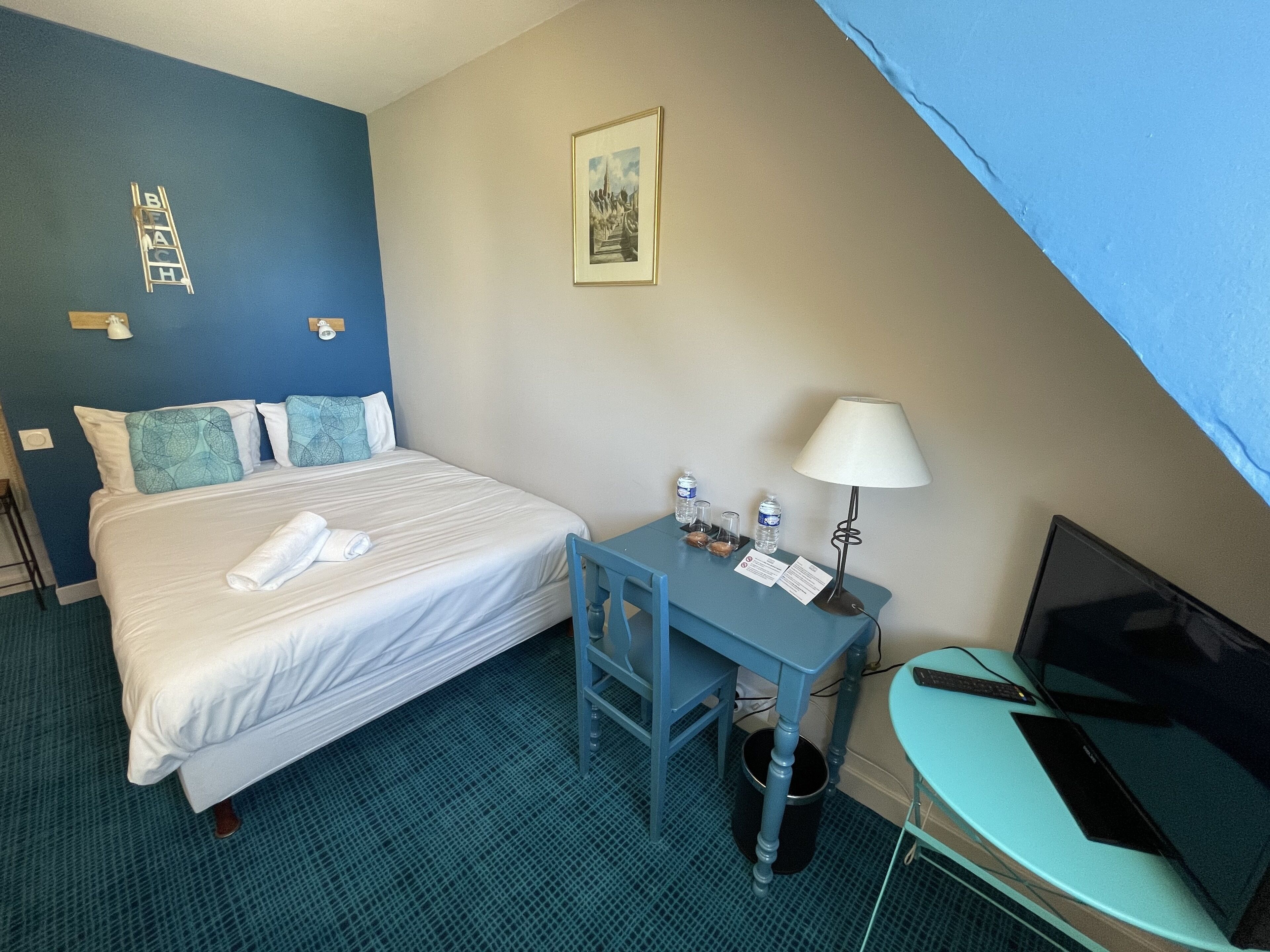 Una habitación de hotel con una cama, un escritorio con una lámpara, un televisor y un cuadro en la pared.