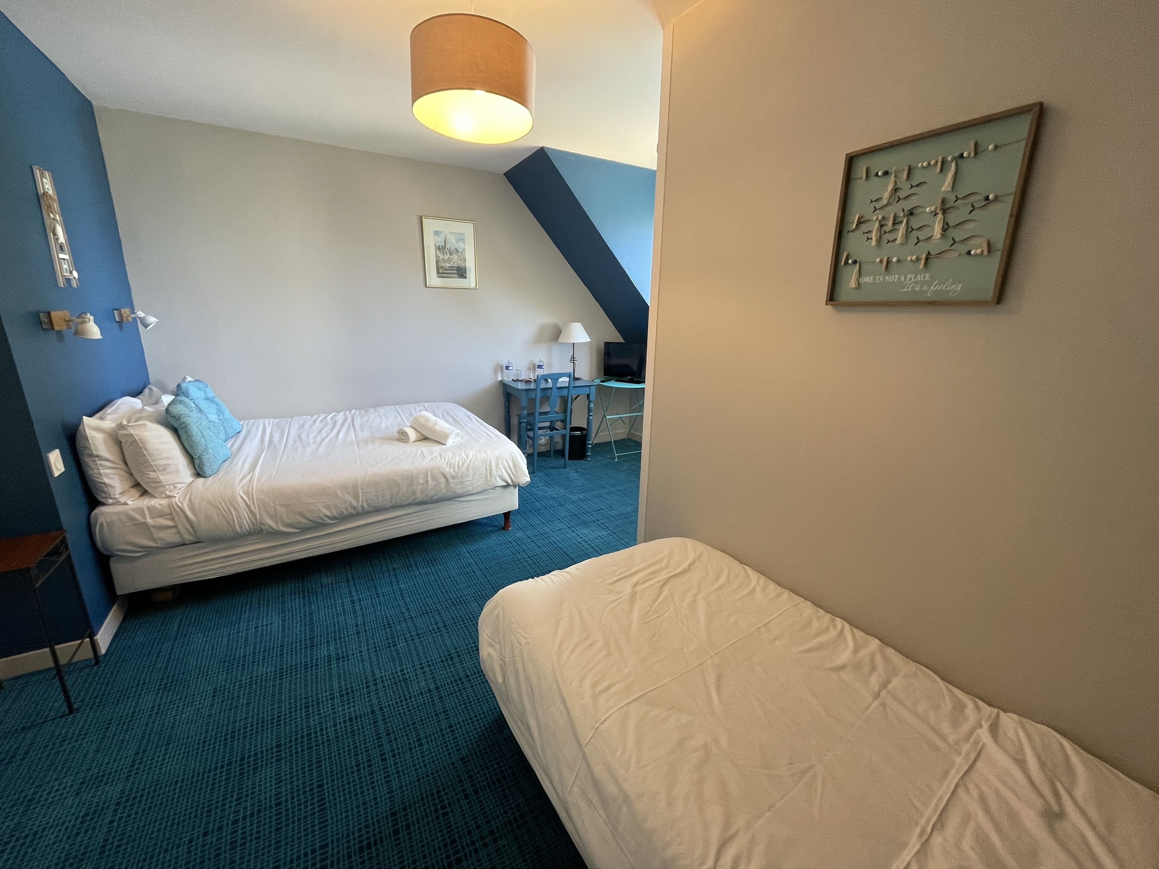 A hotel room with a bed, a desk, and a chair. There is a lamp on the desk and a framed quote on the wall.