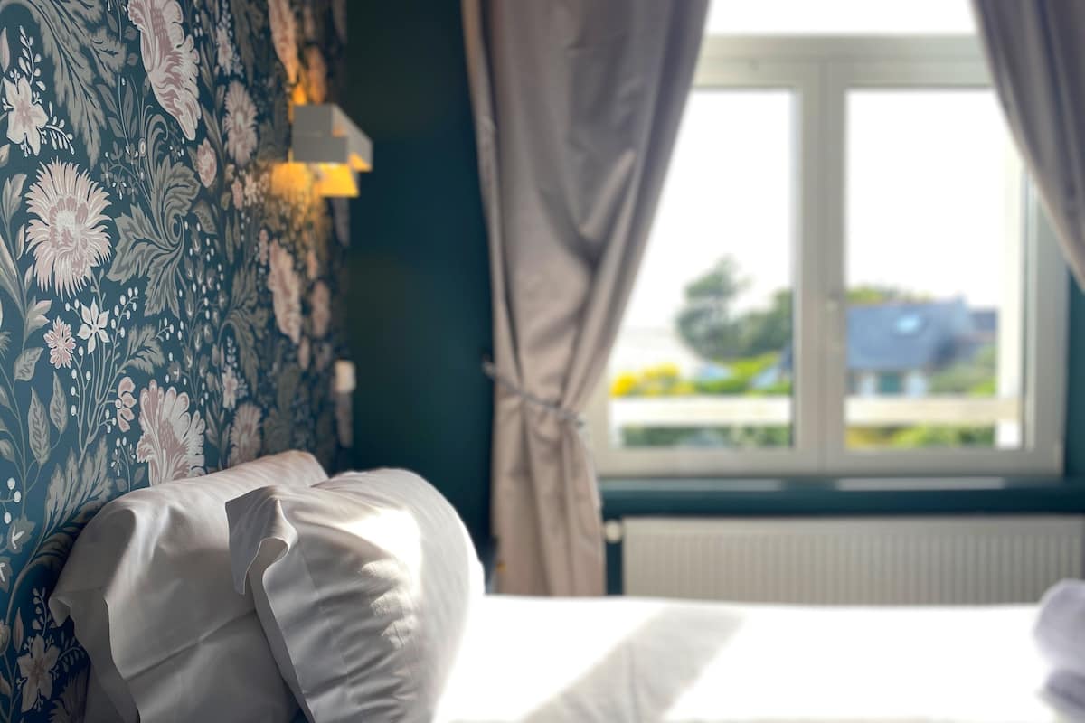 A neatly made bed with white linens, a floral-patterned wall, and a window with curtains.