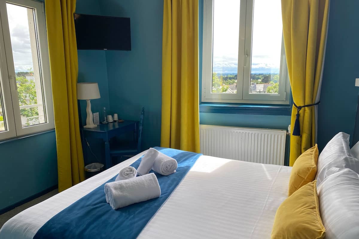 A hotel room with a bed, a desk, a chair, a window with curtains, and a view of the landscape.