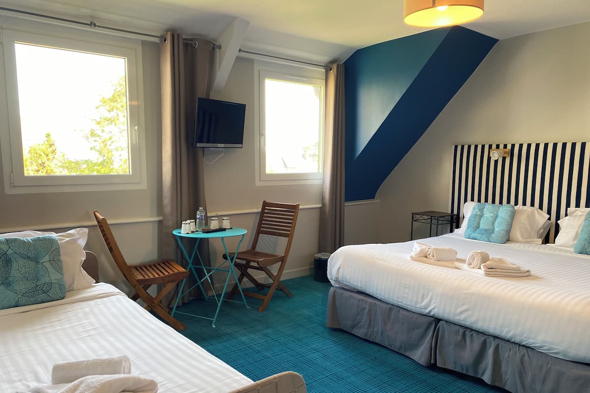 A hotel room with two beds, a small table with chairs, a TV, and a view of trees through the windows.