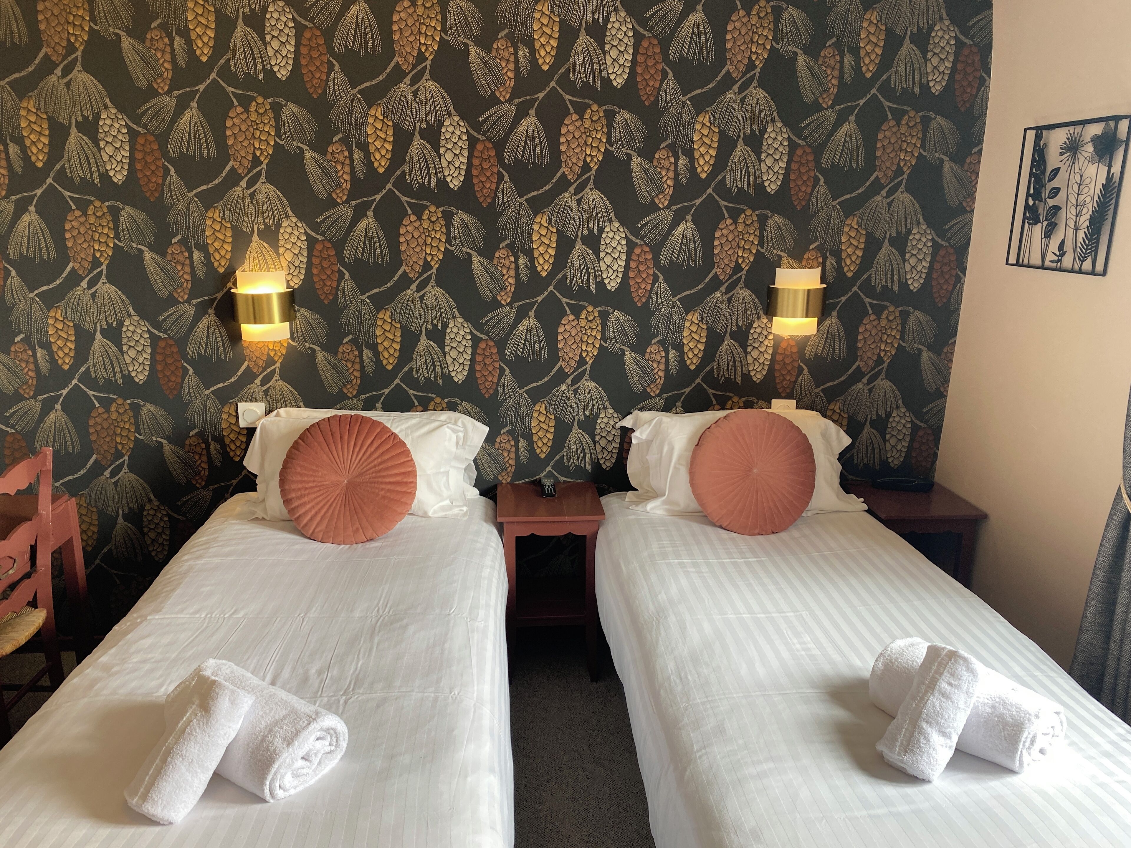 Hotelkamer met twee bedden, elk voorzien van witte lakens en een enkel bruin kussen. Twee nachtkastjes met elk een lamp. De wanden hebben behang met een bladmotief.