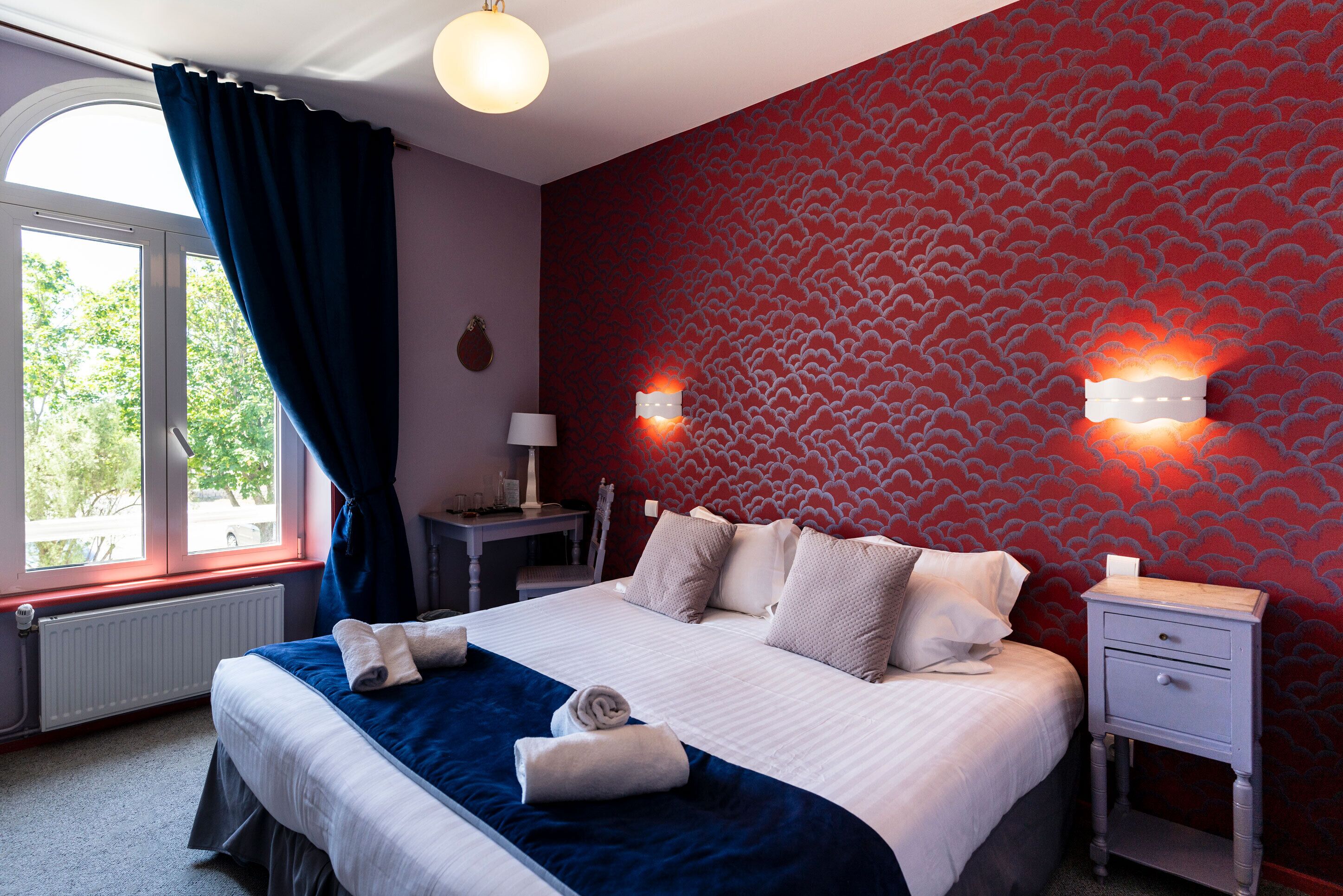 Un lit bien fait, avec des oreillers et une couverture, une table de chevet avec une lampe, et un mur recouvert d’un papier peint à motifs rouges.
