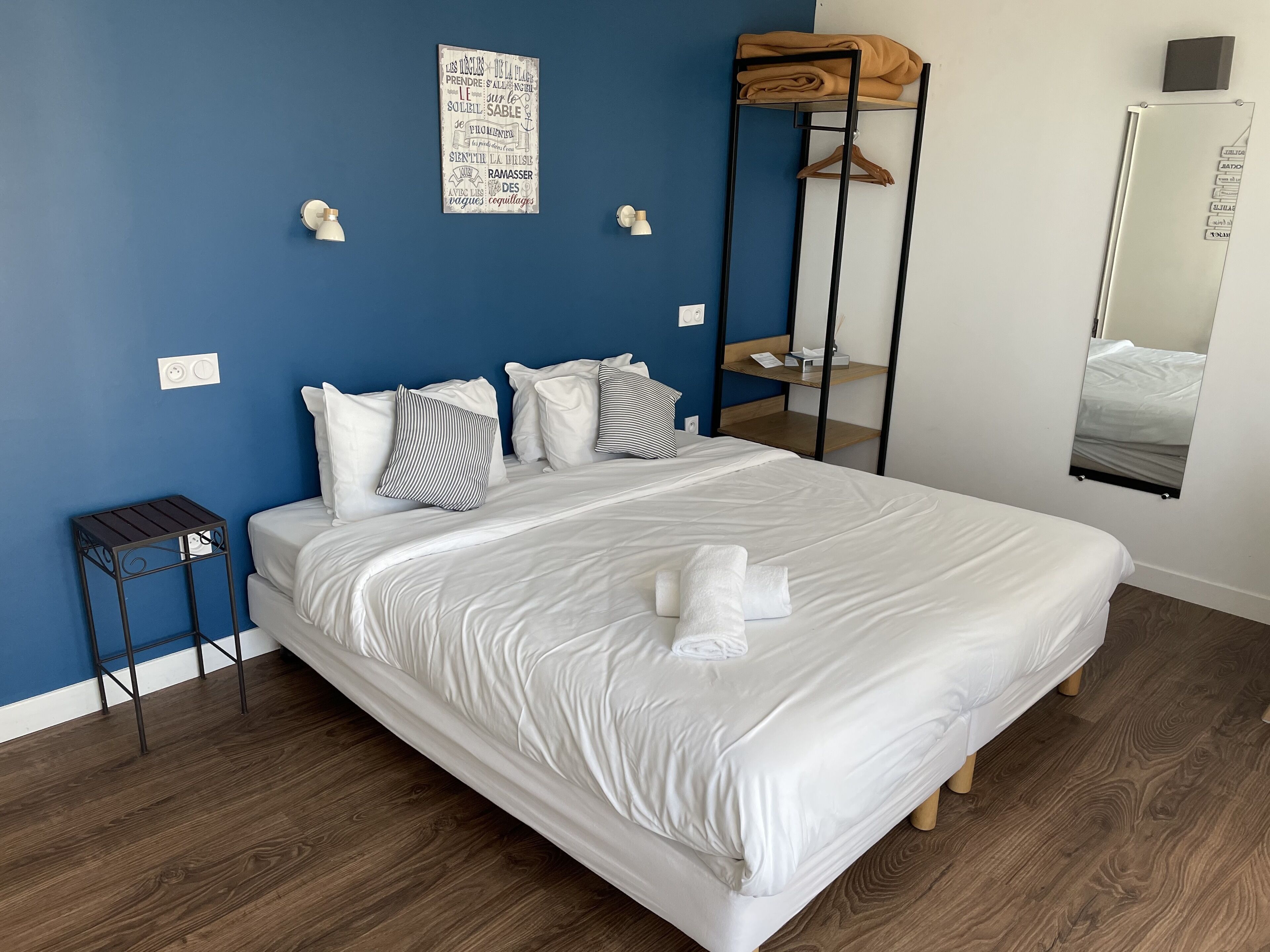 A neatly made bed with white linens, a blue wall, a wooden floor, and a small side table.