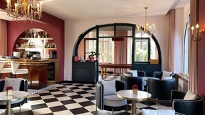 A bar area with a checkered floor, seating with round tables, and a large arched window. - L'Escale Hôtel Bellevue (Tregastel)