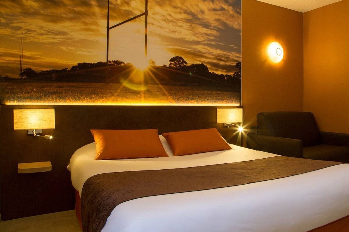 Habitación de hotel con una cama grande, dos lámparas de noche, una silla, una mesita y una luz empotrada en la pared.