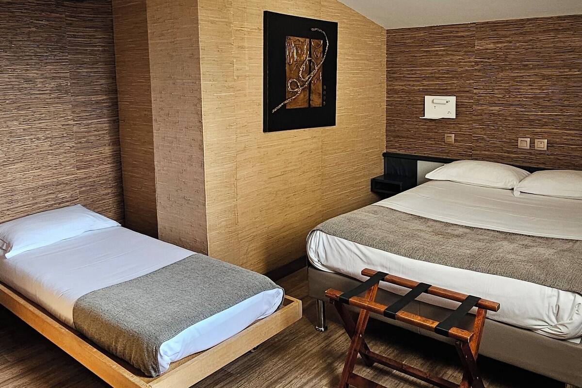 Una habitación con dos camas, un banco de madera, una lámpara en el techo y un cuadro en la pared.