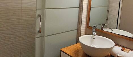 Ein modernes Badezimmer mit einer Duschkabine aus Glas, einem Holzausziehtisch mit weißem Waschbecken und einem großen Spiegel mit Beleuchtung darüber.