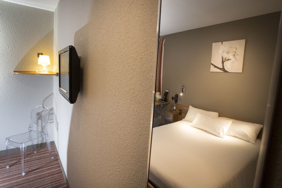 Una habitación de hotel con cama, mesita de noche, lámpara y un cuadro en la pared.