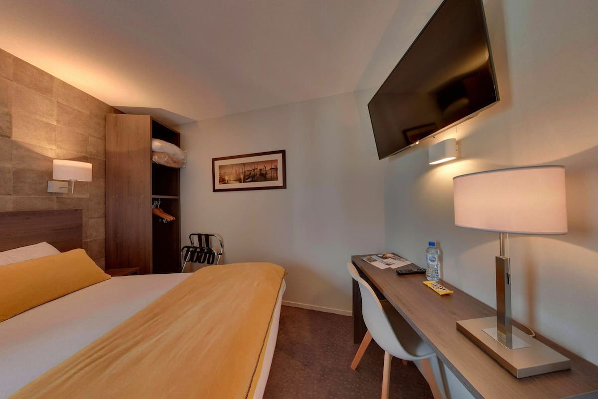 A hotel room with a bed, a desk, a chair, a TV, a lamp, and a picture on the wall.