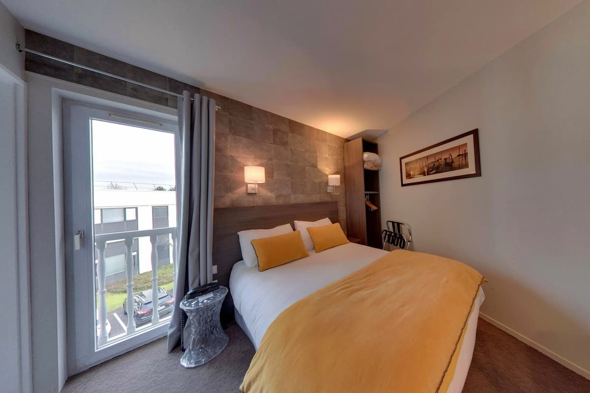 A hotel room with a bed, a wardrobe, a bedside table, a picture on the wall, and a view of the outside.