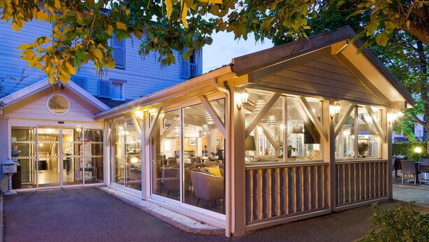 A covered outdoor seating area with glass walls, a wooden deck, and a building with a white facade in the background. - Hotel Acadie Villebon-sur-Yvette (Villebon-sur-Yvette)