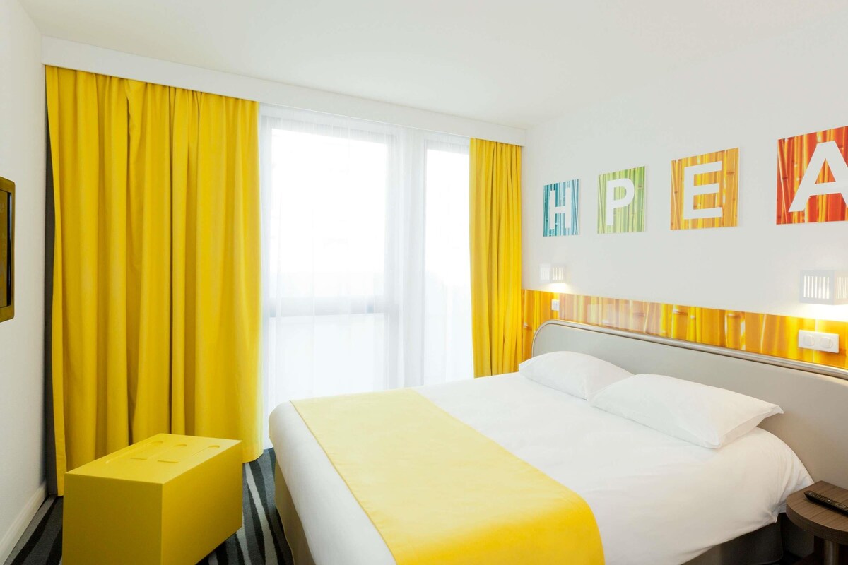 Habitación de hotel con cama, un pequeño taburete amarillo, una mesita de noche y una ventana con cortinas amarillas.